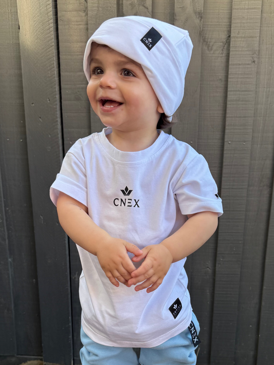 Child wearing a white t-shirt with 'CNEX' logo, light blue pants, and white sneakers standing on a wooden deck.