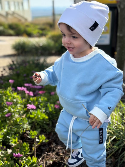 Child in light blue outfit with white beanie standing outdoors near flowers