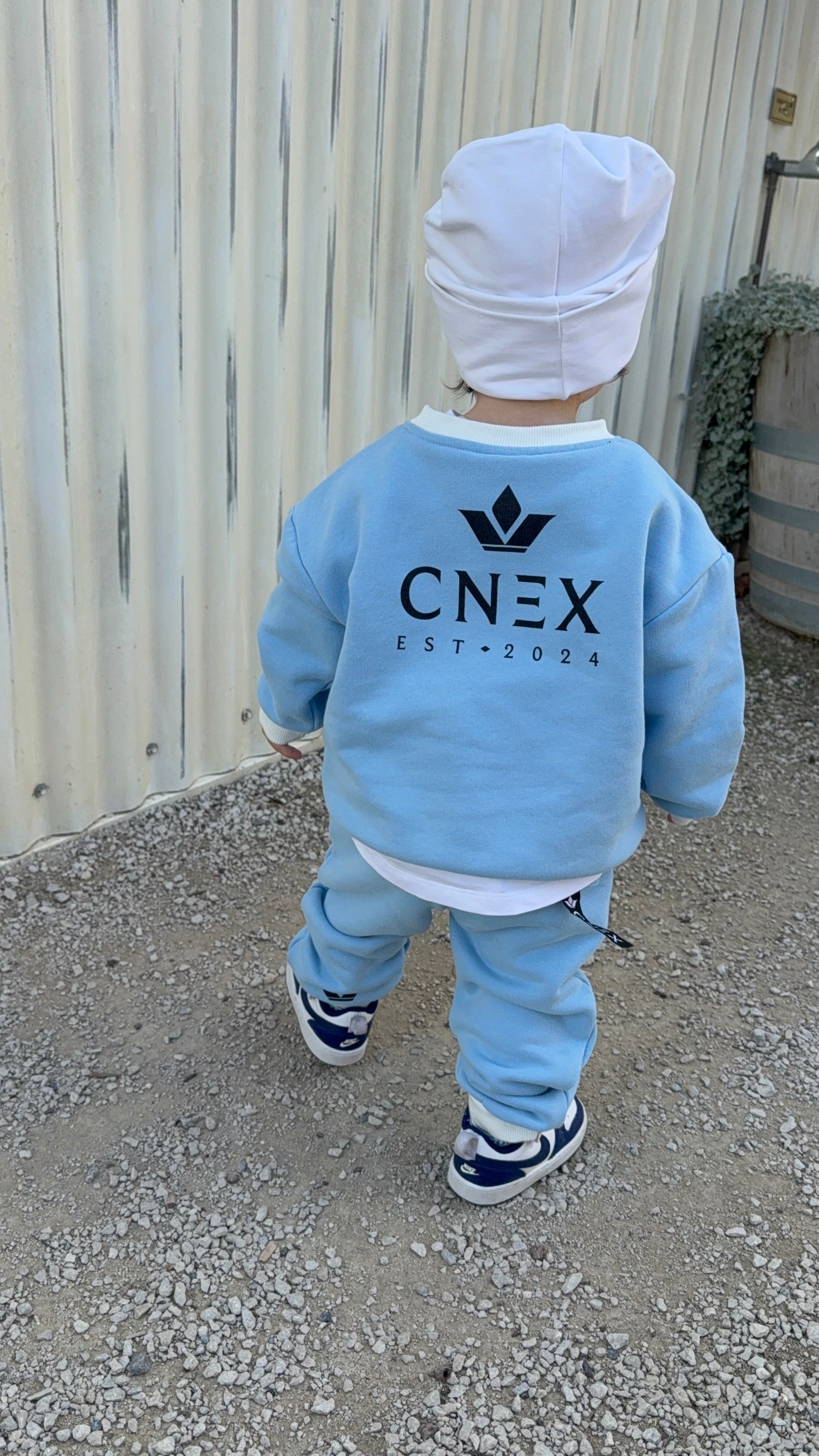 Child wearing a light blue CNEX outfit standing on a gravel surface.